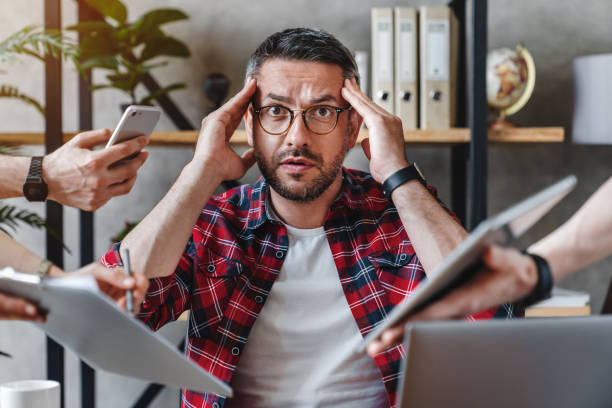 przepracowany biznesmen siedzący przy laptopie przeładowany pracą wielu zadań w nowoczesnym biurze - hard life zdjęcia i obrazy z banku zdjęć