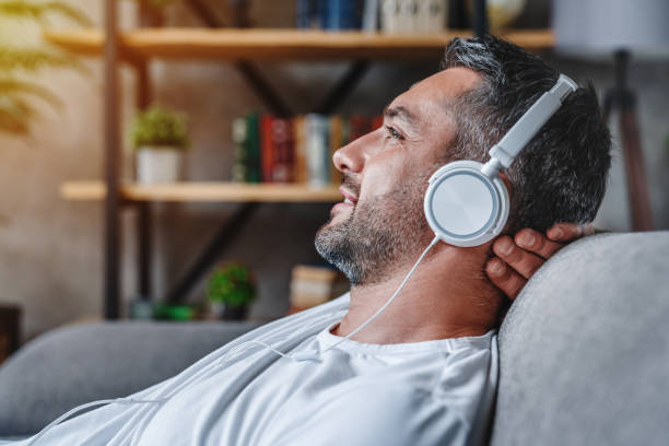 middle aged man listening music with headphones relaxed in sofa at his home - ouvir musica imagens e fotografias de stock