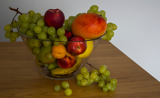 Colorful fresh fruits in transparent glass vase on  textured wooden table. Juicy grape, nectarines and pears on kitchen table. Light grey background with copy space