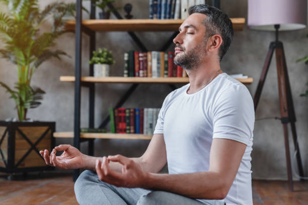 Middle aged man meditating at his living room floor sitting Middle aged man meditating at his living room floor sitting grey hair on floor stock pictures, royalty-free photos & images
