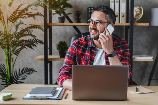 portret uśmiechniętego siwego mężczyzny z brodą siedzącego przednie stołem z laptopem, rozmawiającego przez telefon - old human face men ceo zdjęcia i obrazy z banku zdjęć