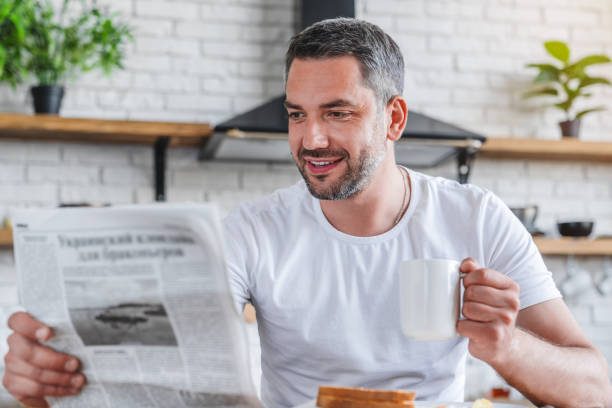 Young adult caucasian man reading newspaper while having breakfast in modern kitchen Young adult caucasian man reading newspaper while having breakfast in modern kitchen reading newspaper stock pictures, royalty-free photos & images