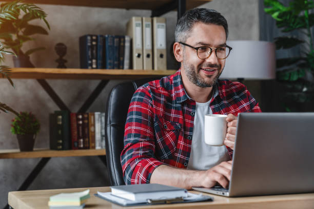 istock Mid adult man using laptop on desk at home and drinking coffee 1253834484
