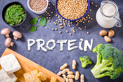 Top view of various kinds of vegan protein sources like tofu, tempeh, soy beans, soy milk, mushrooms, wakame, lentils, peanuts, spinach and chick peas. All the objects are at the borders of the image and at the center is written \