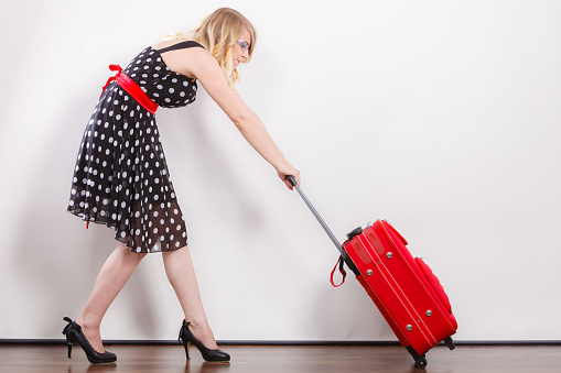 Traveling vacation concept. Elegant young woman in full length wearing polka dot black dress pulling heavy red travel bag
