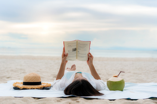 Young Asian woman lying on a tropical summer beach sea for relax reading book with coconut juice beside her. Summer, holidays, vacation and happy people in Thailand concept.