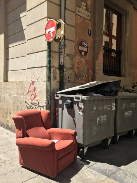 Old armchair next to industrial garbage containers Valencia, Spain - June 12, 2020: Old armchair sitting next to garbage containers in the street. People leave them like this hoping the garbage collectors or someone needing it will take it overflowing trash dumpster stock pictures, royalty-free photos & images