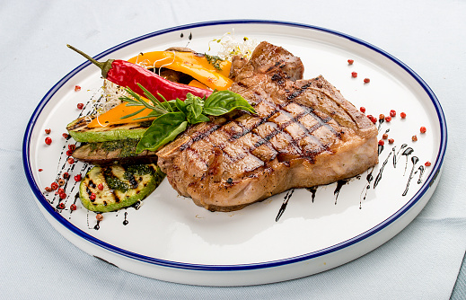 Tender grilled steak or T-bone steak served with grilled vegetables on a white plate.