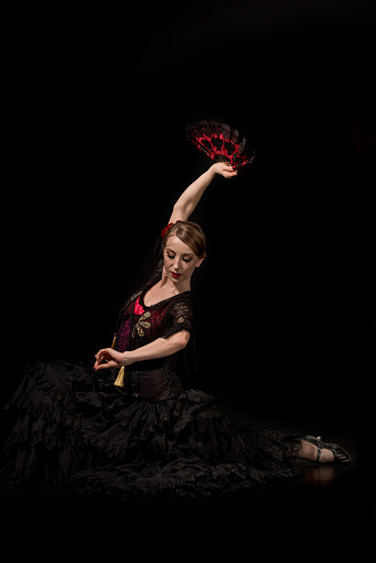 elegant flamenco dancer holding fan above head and sitting on black