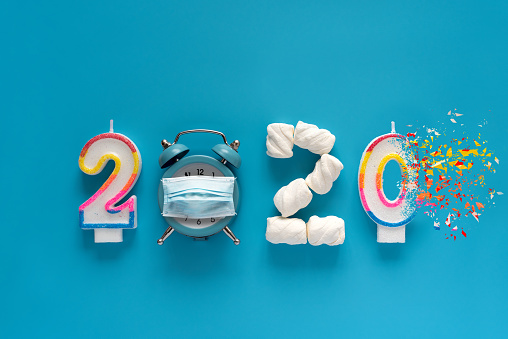 2020 year made of candles, marshmallow and alarm clock wearing in protective mask with dispersion effect on blue background. New Year 2021 celebration or end of year 2020 concept. Top view. Flat lay