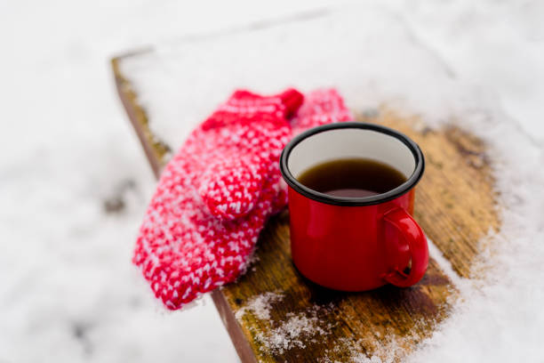 Red mug with hot coffee or tea drink on snow in winter. Red knitted mittens on table