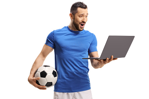 Excited soccer player holding a ball and a looking at a laptop computer isolated on white background