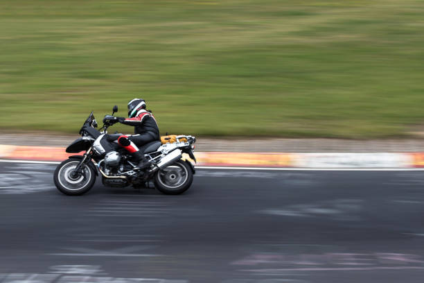 A handsome young adult male is riding a red vintage Cafe Racer styled Motorcycle around a bend wearing an open faced helmet, sunglasses and gloves. -- Post processing to look Vintage.  The man is wearing all black and the sun is low enough to cast alluring shadows.
