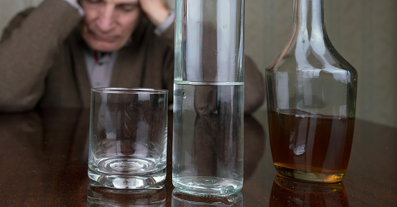 disappointed old man holds hands on head sitting at brown wooden table with bottles and glass at home