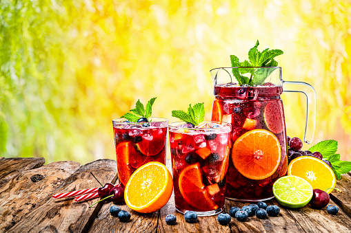 Refreshing drink for summer: pitcher and two glasses of cold refreshing sangria with fruits shot on rustic wooden table against yellow lush foliage background. Fruits included in the composition are grape, orange, lime and berries. The sangria glasses are garnished with mint leaves. Two red and white drinking straws complete the composition. Predominant colors are red, brown and yellow. High resolution studio digital capture taken with Sony A7rII and Sony FE 90mm f2.8 macro G OSS lens