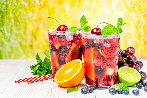 Refreshing drink for summer: two glasses of cold refreshing sangria with fruits shot on white table against yellow lush foliage background. Fruits included in the composition are grape, orange, lime and berries. The sangria glasses are garnished with mint leaves. Two red and white drinking straws complete the composition. Predominant colors are red and yellow. High resolution studio digital capture taken with Sony A7rII and Sony FE 90mm f2.8 macro G OSS lens