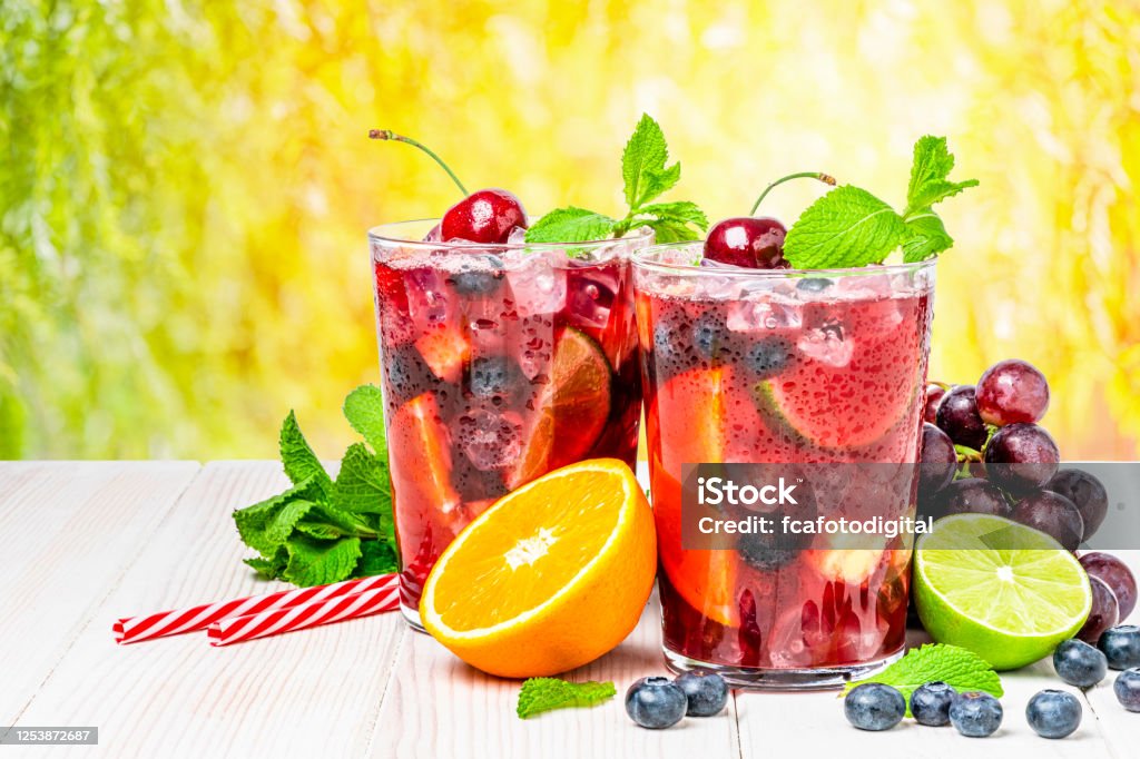 Cold refreshing sangria with fruits on rustic wooden table Refreshing drink for summer: two glasses of cold refreshing sangria with fruits shot on white table against yellow lush foliage background. Fruits included in the composition are grape, orange, lime and berries. The sangria glasses are garnished with mint leaves. Two red and white drinking straws complete the composition. Predominant colors are red and yellow. High resolution studio digital capture taken with Sony A7rII and Sony FE 90mm f2.8 macro G OSS lens Sangria Stock Photo
