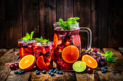 Refreshing drink for summer: pitcher and two glasses of cold refreshing sangria with fruits shot on rustic wooden table. Fruits included in the composition are grape, orange, lime and berries. The sangria glasses are garnished with mint leaves. Two red and white drinking straws complete the composition. Predominant colors are red and brown. High resolution 42Mp studio digital capture taken with Sony A7rII and Sony FE 90mm f2.8 macro G OSS lens