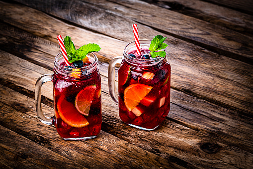 Refreshing drink for summer: high angle view of two Mason jars of cold refreshing sangria shot on rustic wooden table. The sangria glasses are garnished with mint leaves. Two red and white drinking straws complete the composition. Predominant colors are red and brown. High resolution 42Mp studio digital capture taken with Sony A7rII and Sony FE 90mm f2.8 macro G OSS lens