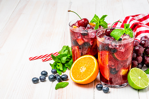 Refreshing drink for summer: two glasses of cold refreshing sangria with fruits arranged all around the glasses shot on white table. Fruits included in the composition are grape, orange, lime and berries. The sangria glasses are garnished with mint leaves. Two red and white drinking straws complete the composition. The composition is at the right of an horizontal frame leaving useful copy space for text and/or logo at the left. Predominant colors are red and white. High resolution 42Mp studio digital capture taken with Sony A7rII and Sony FE 90mm f2.8 macro G OSS lens