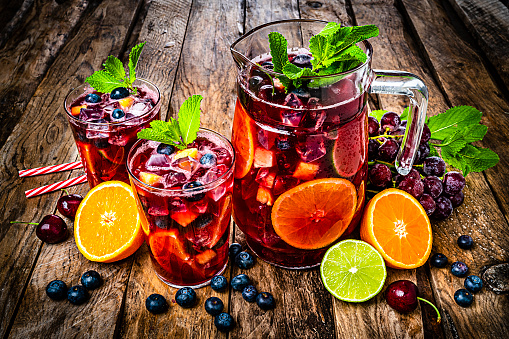 Refreshing drink for summer: high angle view of a pitcher and two glasses of cold refreshing sangria with fruits shot on rustic wooden table. Fruits included in the composition are grape, orange, lime and berries. The sangria glasses are garnished with mint leaves. Two red and white drinking straws complete the composition. Predominant colors are red and brown. High resolution 42Mp studio digital capture taken with Sony A7rII and Sony FE 90mm f2.8 macro G OSS lens