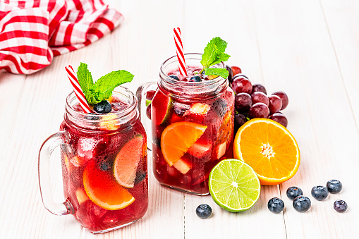 Refreshing drink for summer: high angle view of two Mason jars of cold refreshing sangria with fruits arranged all around the jars shot on white table. Fruits included in the composition are grape, orange, lime and berries. The sangria glasses are garnished with mint leaves. Two red and white drinking straws complete the composition. Predominant colors are red and white. High resolution 42Mp studio digital capture taken with Sony A7rII and Sony FE 90mm f2.8 macro G OSS lens