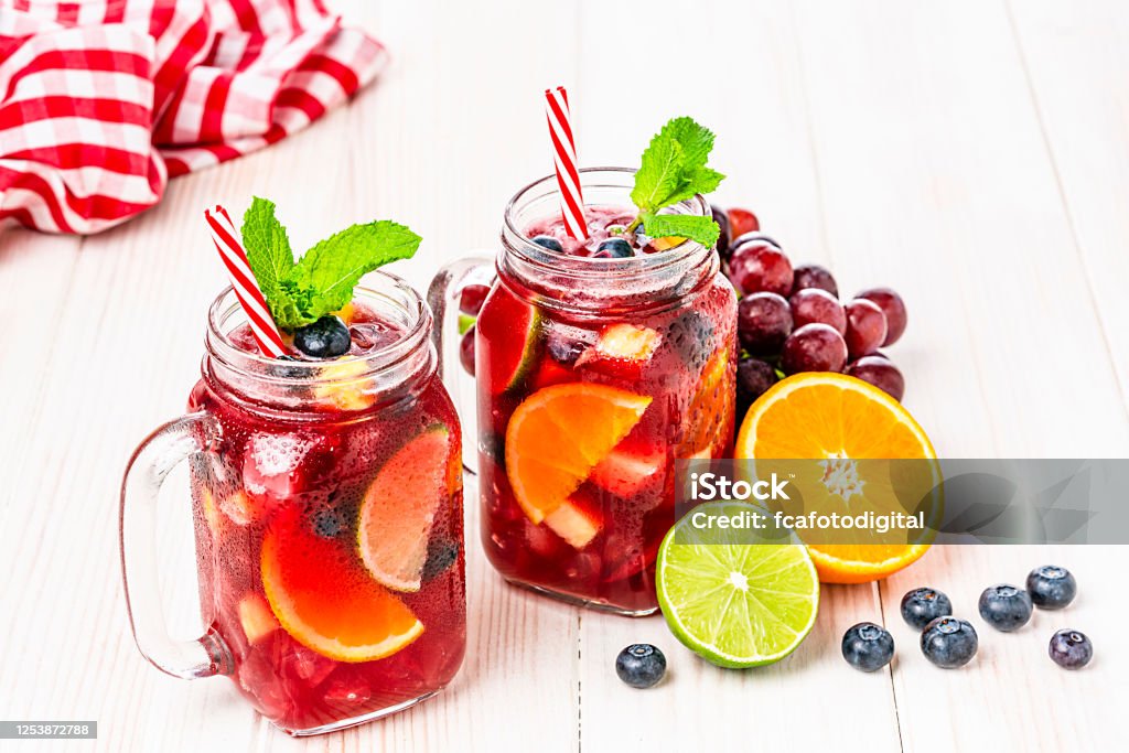 Two Mason jars of cold refreshing sangria with fruits on white table Refreshing drink for summer: high angle view of two Mason jars of cold refreshing sangria with fruits arranged all around the jars shot on white table. Fruits included in the composition are grape, orange, lime and berries. The sangria glasses are garnished with mint leaves. Two red and white drinking straws complete the composition. Predominant colors are red and white. High resolution 42Mp studio digital capture taken with Sony A7rII and Sony FE 90mm f2.8 macro G OSS lens Sangria Stock Photo
