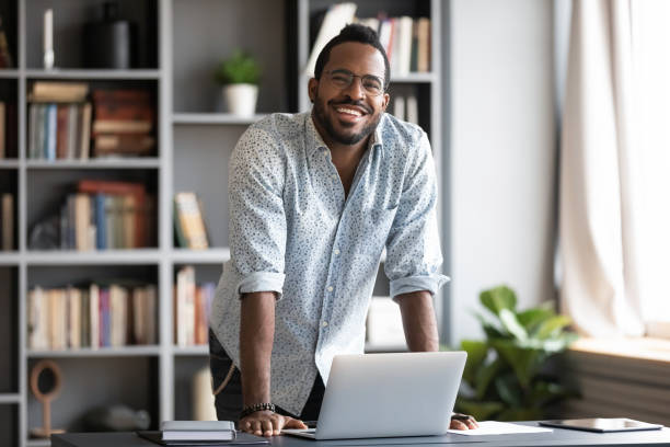 hombre africano de pie se inclinó sobre el ordenador portátil sonriendo mirando a la cámara - business looking at camera office new fotografías e imágenes de stock
