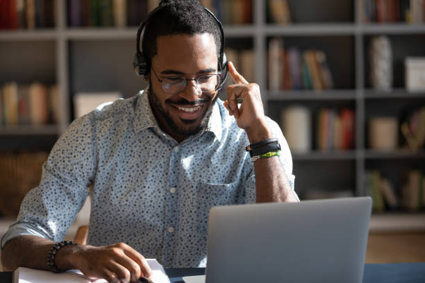 African man wear headset watching webinar video course Focused African man sitting at desk wear headset watching webinar video course gain new knowledge use on-line application website. Manager talk to client provide professional help and support concept online skills stock pictures, royalty-free photos & images
