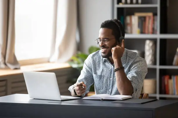 Photo of African guy learn online wearing headset looking at laptop screen