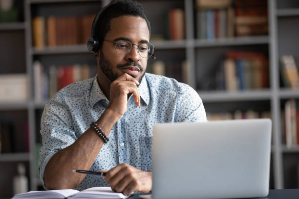 African guy wearing headset learn foreign language using internet websites Focused African man wear headphones with microphone looking at laptop screen listens audio lesson learn foreign language with tutor makes video call. Student watching webinar, e study on-line concept virtual college education stock pictures, royalty-free photos & images