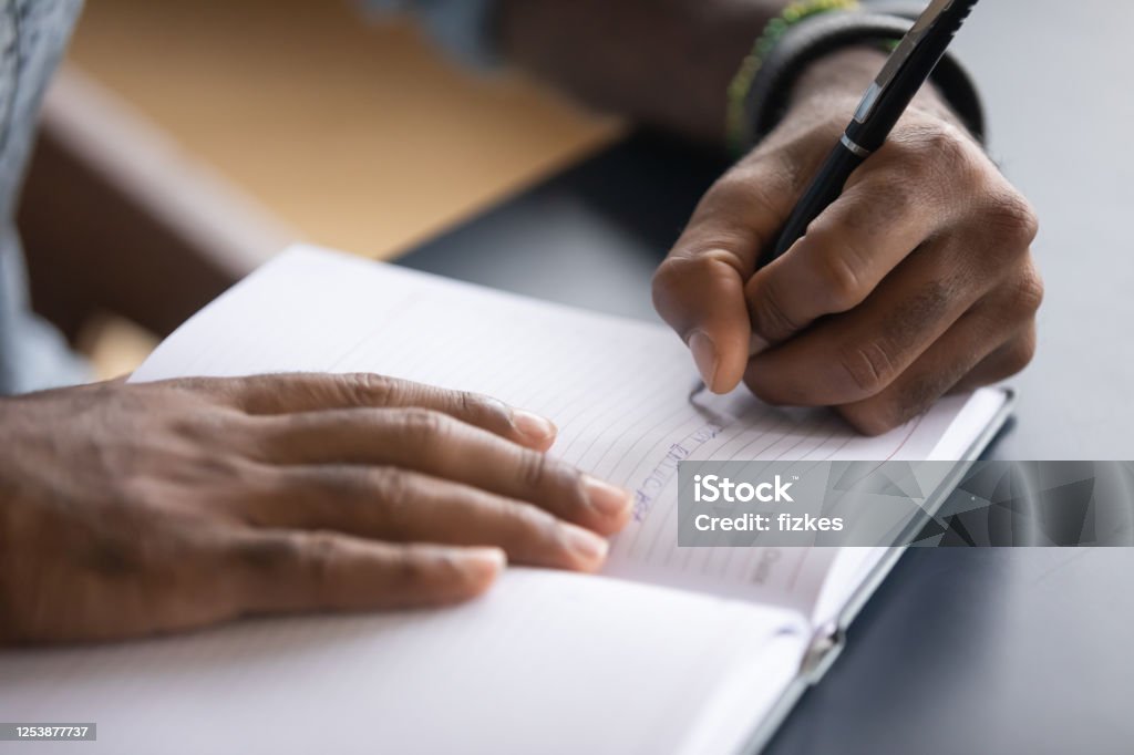 African man seated at desk writes information on notebook closeup African man sit at desk hold pen keeps startup business ideas, plans, creative thoughts to notebook close up image. Makes appointment notes time, writes important things and to-do list not to forget Writing - Activity Stock Photo