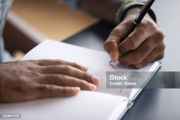 African Man Seated At Desk Writes Information On Notebook Closeup Stock Photo - Download Image Now