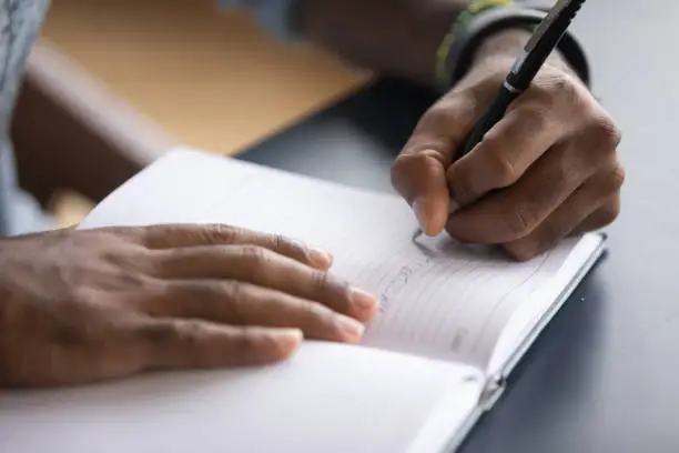 African man sit at desk hold pen keeps startup business ideas, plans, creative thoughts to notebook close up image. Makes appointment notes time, writes important things and to-do list not to forget