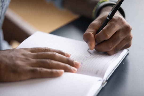 african man seated at desk writes information on notebook closeup - capital letter fotos imagens e fotografias de stock