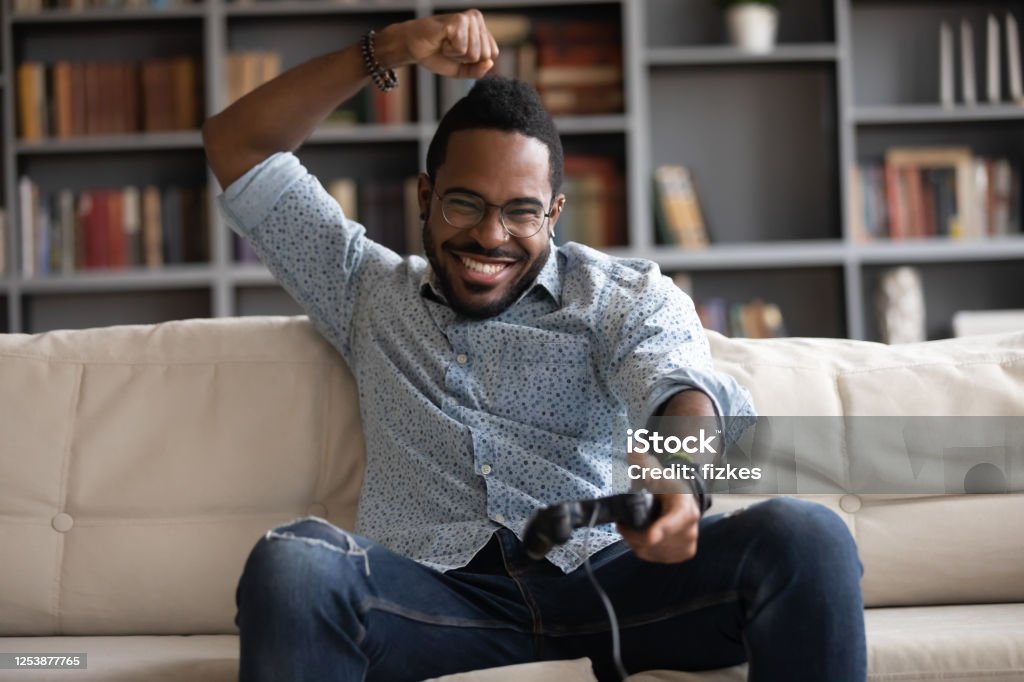 Excited African guy holding joypad celebrating victory in playstation games Excited successful african guy sit on couch in living room spend weekend free time at home holding joypad celebrating victory. Having fun using modern technology devices virtual video games concept Video Game Stock Photo