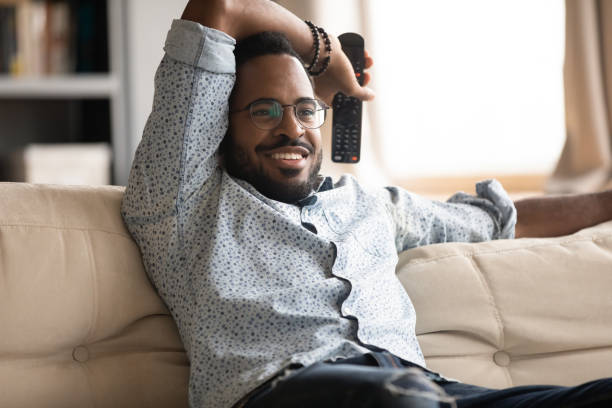 African guy resting on couch holding remote control watching tv Relaxed African fellow resting on couch holding remote control watching tv shows television programs or favourite series movie, football fan enjoy match sport game, weekend lazy day at home concept hand fan photos stock pictures, royalty-free photos & images