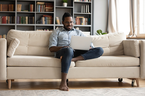 African guy rest on sofa put notebook on lap looking at screen watching movie enjoy lazy free day, creates blog, booking on-line, buyer choose goods makes order, shopping in internet lifestyle concept