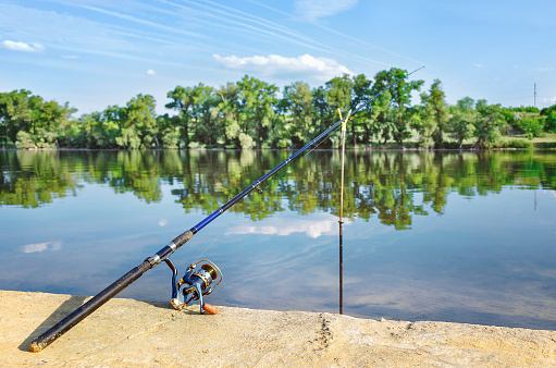 Fishing rod throwed on the river  banks.  Summer sunny day, scenic view of the horizon with trees, reflection of the sky in the water. Concept of countryside getaway.