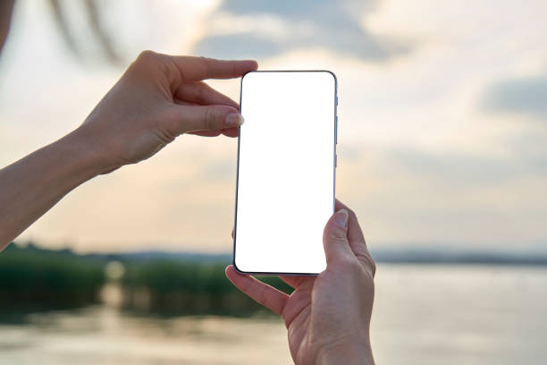 istock Woman's hands holding a smartphone with white screen mockup on blur background during sunset on the lake. 1253882969
