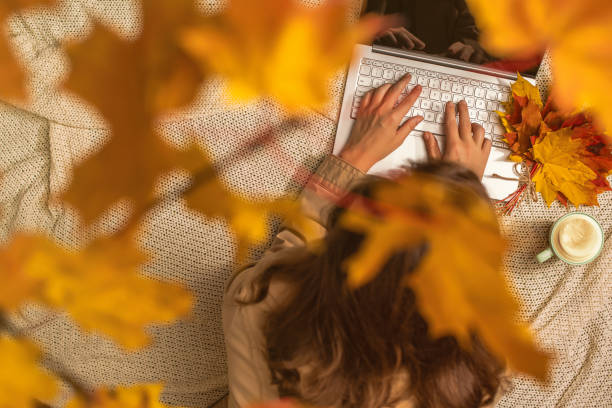 Photo of Autumn. Woman freelancer on the plaid with laptop, cup of coffee under branch of golden maple leaf on the green grass in the park. Concept. Top view