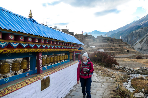 A woman walking along a wall full of prayer wheels on Annapurna Circuit Trek in Himalayas, Nepal. The woman enjoys the trek. There is a high Himalayan chain in the back. Spirituality and meditation