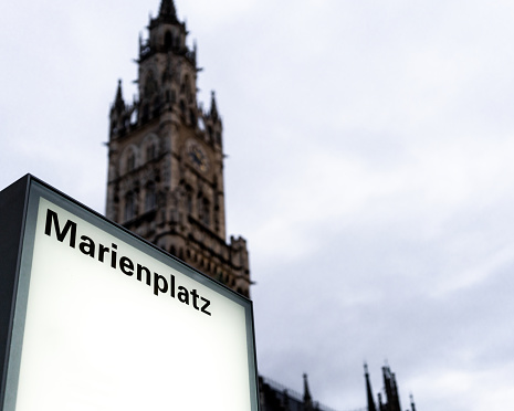 An illuminated modern sign for the Marienplatz town square, in the heart of Munich, Bavaria, with the famous spire of the city's New Town Hall in the background.