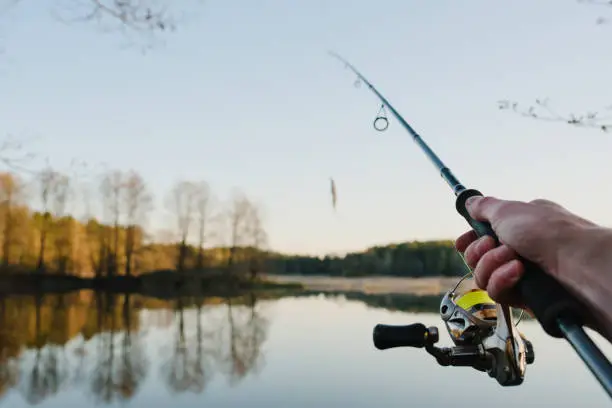 Photo of Fisherman with rod throws bait into the water on river bank. Fishing for pike, perch. Background wild nature. Concept of rural getaway. Man catching a fish on lake or pond with text space. Fishing day.