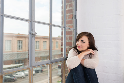 Young woman with a pensive expression sitting on a windowsill overlooking a street in town staring thoughtfully at the scene below