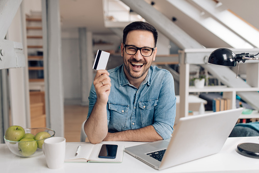 Portrait of a young man holding a credit card and shopping online at his home office.