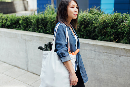 Beautiful young Asian woman walking with reusable shopping bag after grocery shopping.