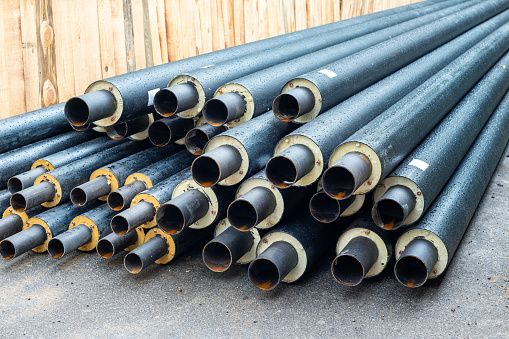 Heap of many new black insulated steel pipes at municipal construction site outdoors. Heating main district pipeline renewal or reconstruction. City development building industrial background.