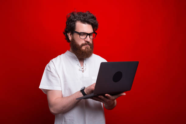 深刻なひげを生やした男は、赤い背景の上にラップトップを使用しています。 - beautiful business lifestyles studio shot ストックフォトと画像