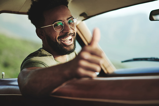 Portrait of a young man showing thumbs up while sitting in a car during a road trip